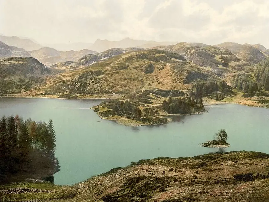 A still tarn ringed by trees with bare fells rising behind it.