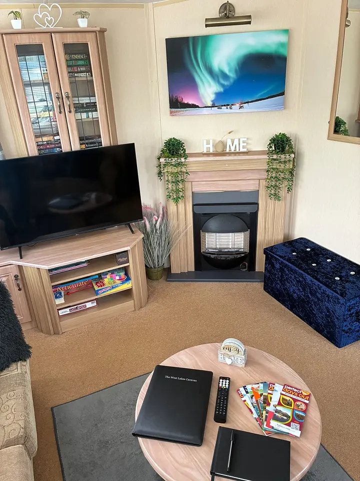 The lounge in The West Lakes Caravan with an electric fire, a framed aurora print above the mantel and board games stacked under the television.